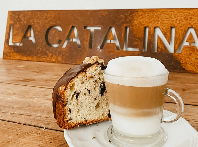 Taza de capuccino con espuma de leche acompañado de una porción de budín de chocolate y frutos secos